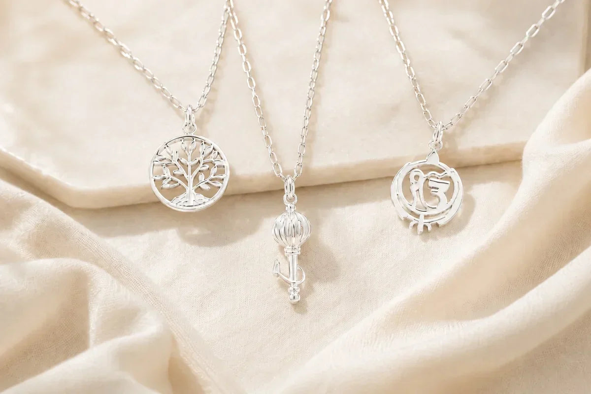Three 92.5 silver pendants on chains — Tree of Life, Vayuputra Gada and Ek Onkar Kripa — laid on cream fabric
